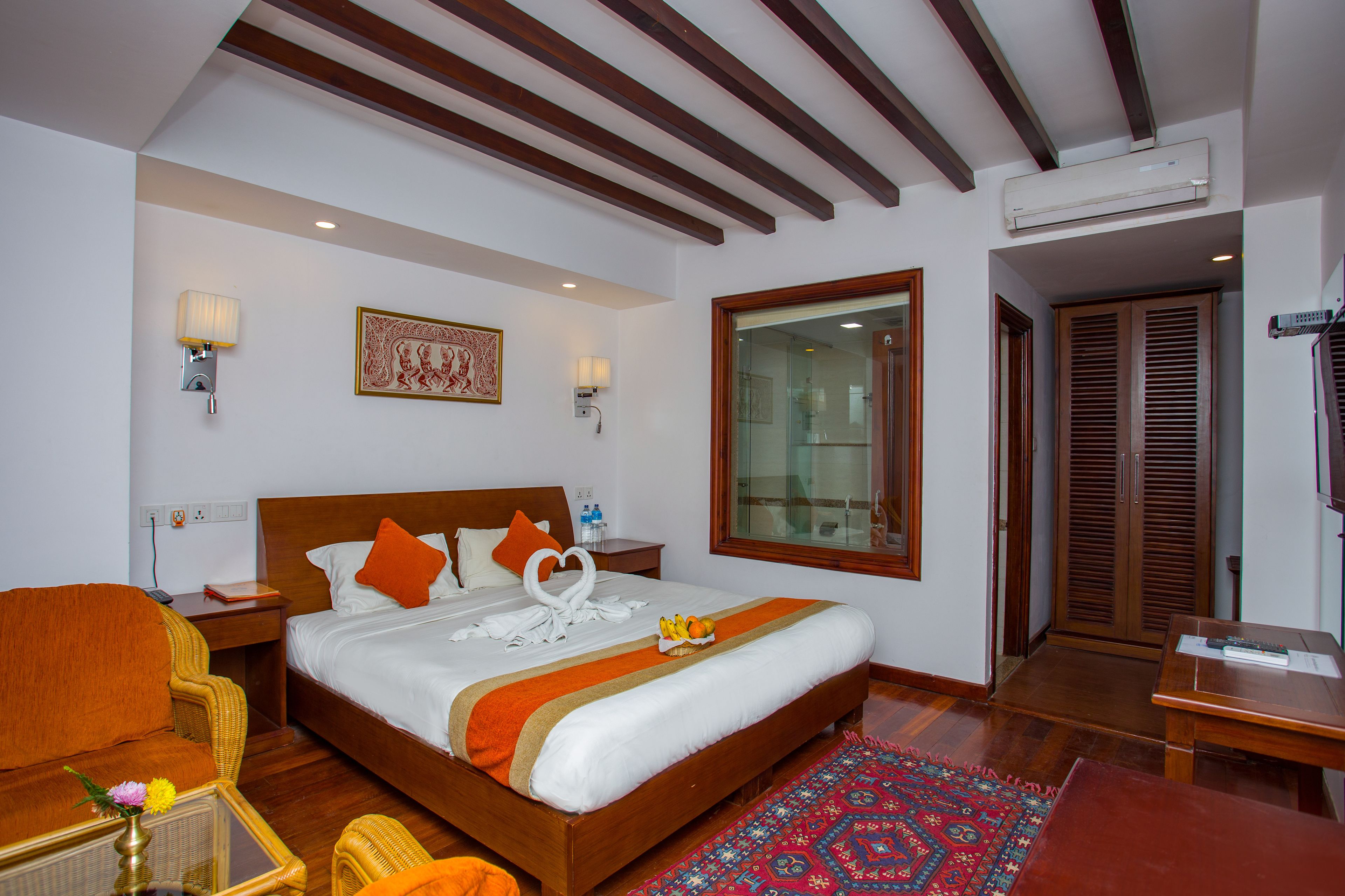 A hotel room with a wooden bed, two bedside lamps, a nightstand, a wicker chair, a small table with a vase, and a red patterned rug.