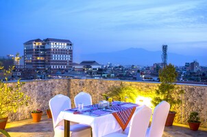 Un espace de restauration aménagé sur le toit, avec une table dressée pour deux personnes, entouré de plantes en pot et offrant une vue sur le paysage urbain, avec des montagnes en arrière-plan.