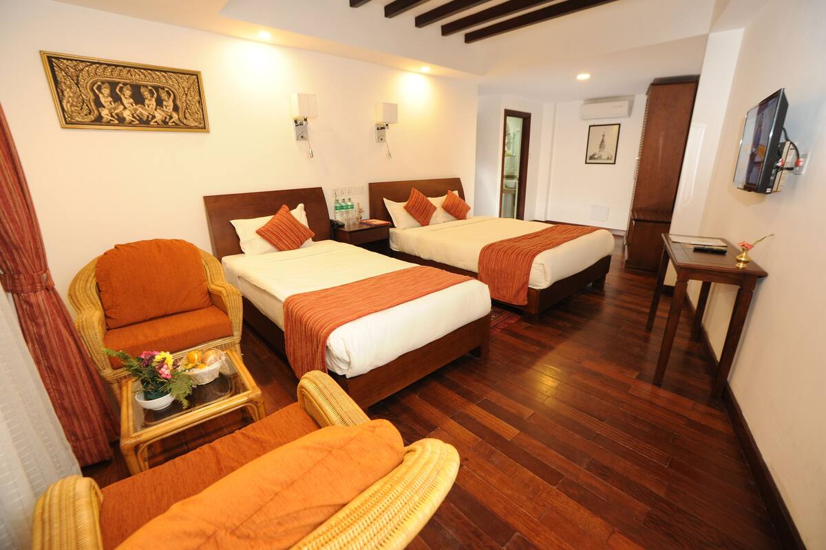 Habitación de hotel con dos camas, una silla, una mesita con un jarrón y un espejo en la pared.