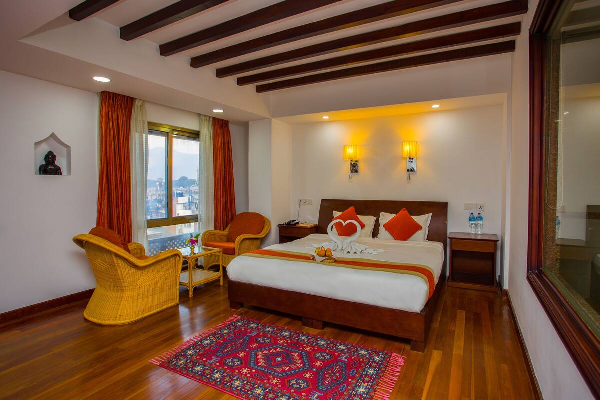 Habitación de hotel con una cama grande, dos lámparas de pared, techo de madera, una silla de mimbre, una mesita pequeña y vista a la ciudad por la ventana.