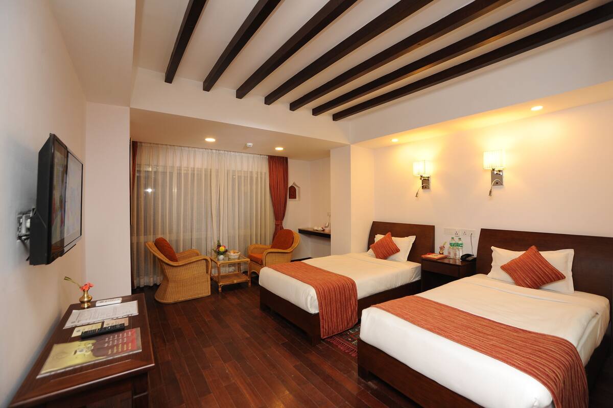 Habitación de hotel con dos camas, piso de madera, televisor, escritorio con silla y ventana con cortinas.