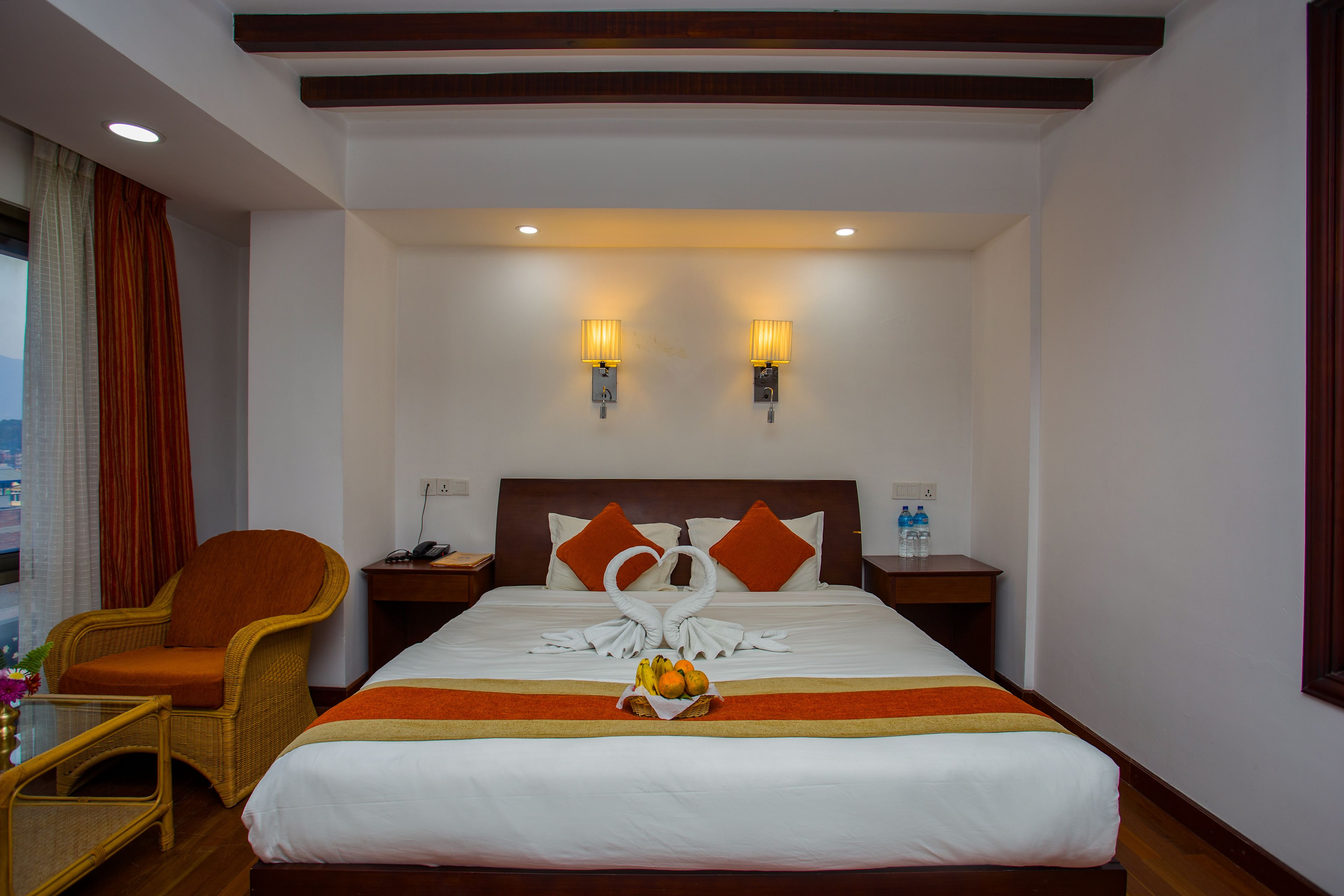 A hotel room with a large bed, two wall-mounted lamps, a wicker chair, a small table with a vase, and a fruit basket on the bed.