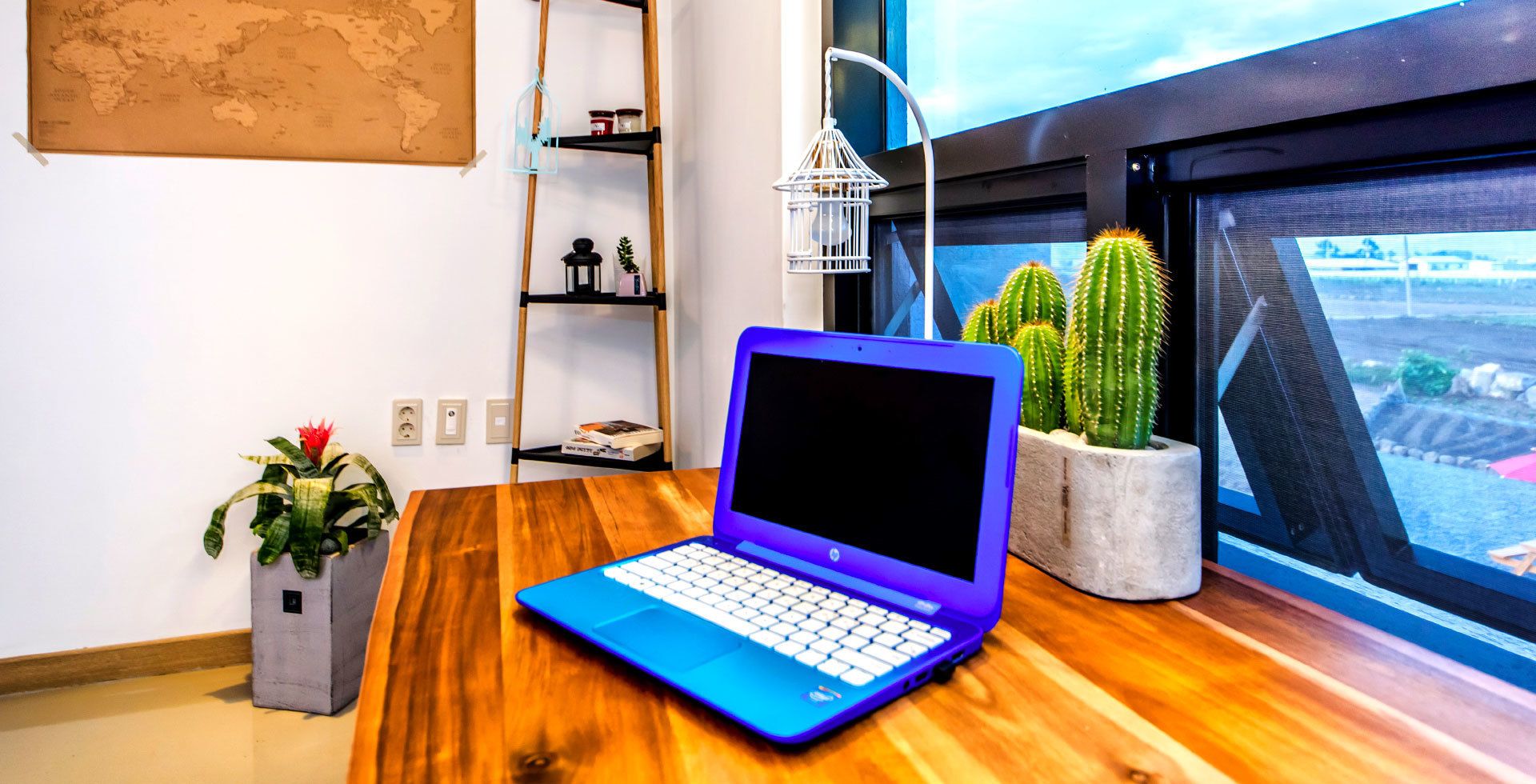 Um laptop sobre uma mesa de madeira, com vista para uma janela, uma planta cacto e um mapa-múndi na parede.