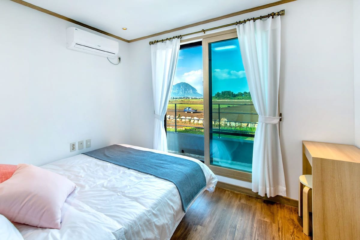 A bedroom with a bed, a bedside table, a window with curtains, and a view of a landscape.