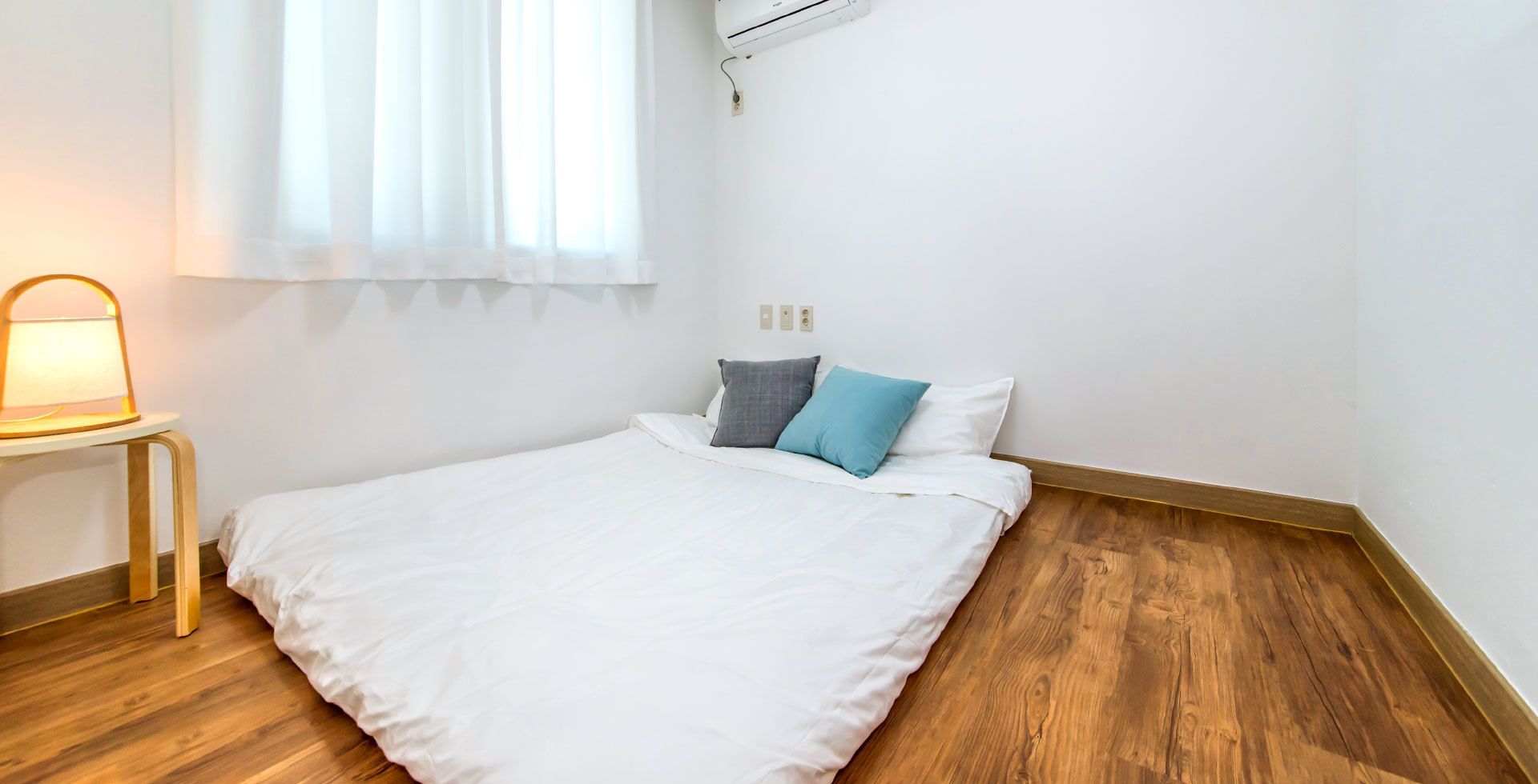 A neatly made bed with white bedding and two pillows, a small bedside table with a lamp, and a wooden floor.