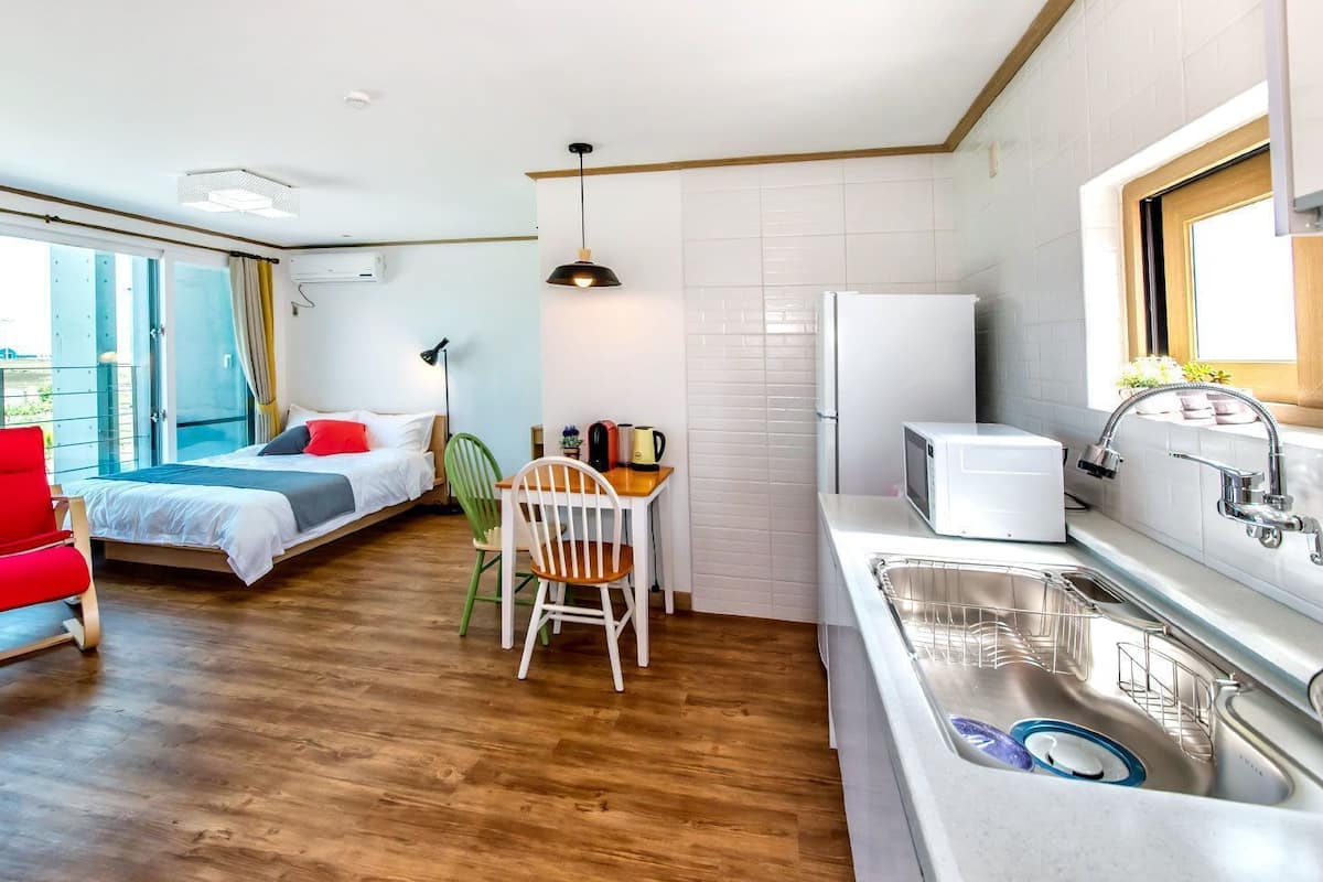A modern room with a bed, a red chair, a dining table, a kitchen with a sink and a microwave, and a large window.