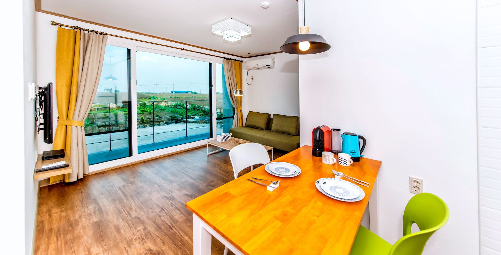 A modern living room with a wooden dining table, a sofa, and a large window with curtains.