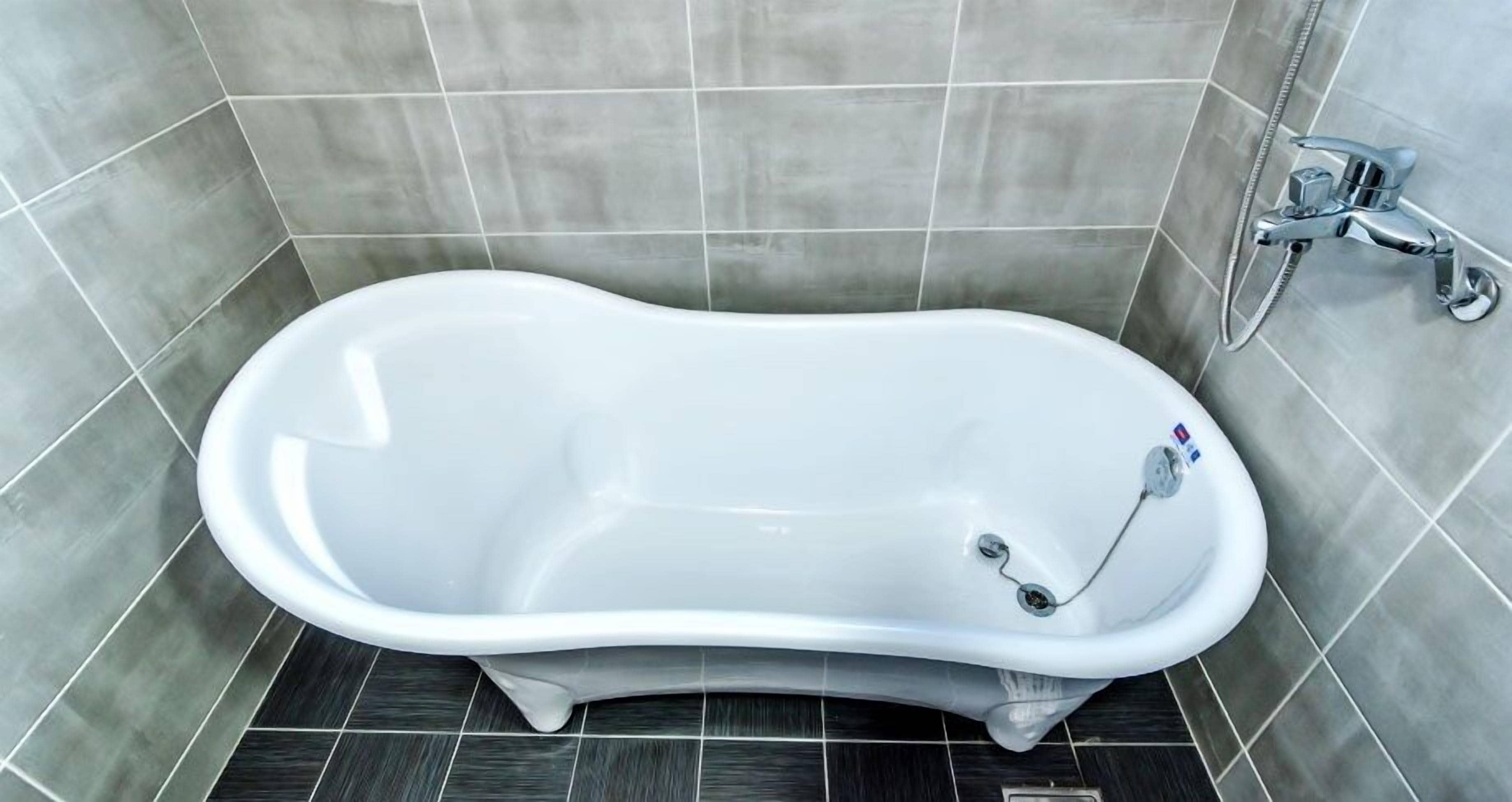 A white freestanding bathtub in a bathroom with grey tiles on the walls and floor.