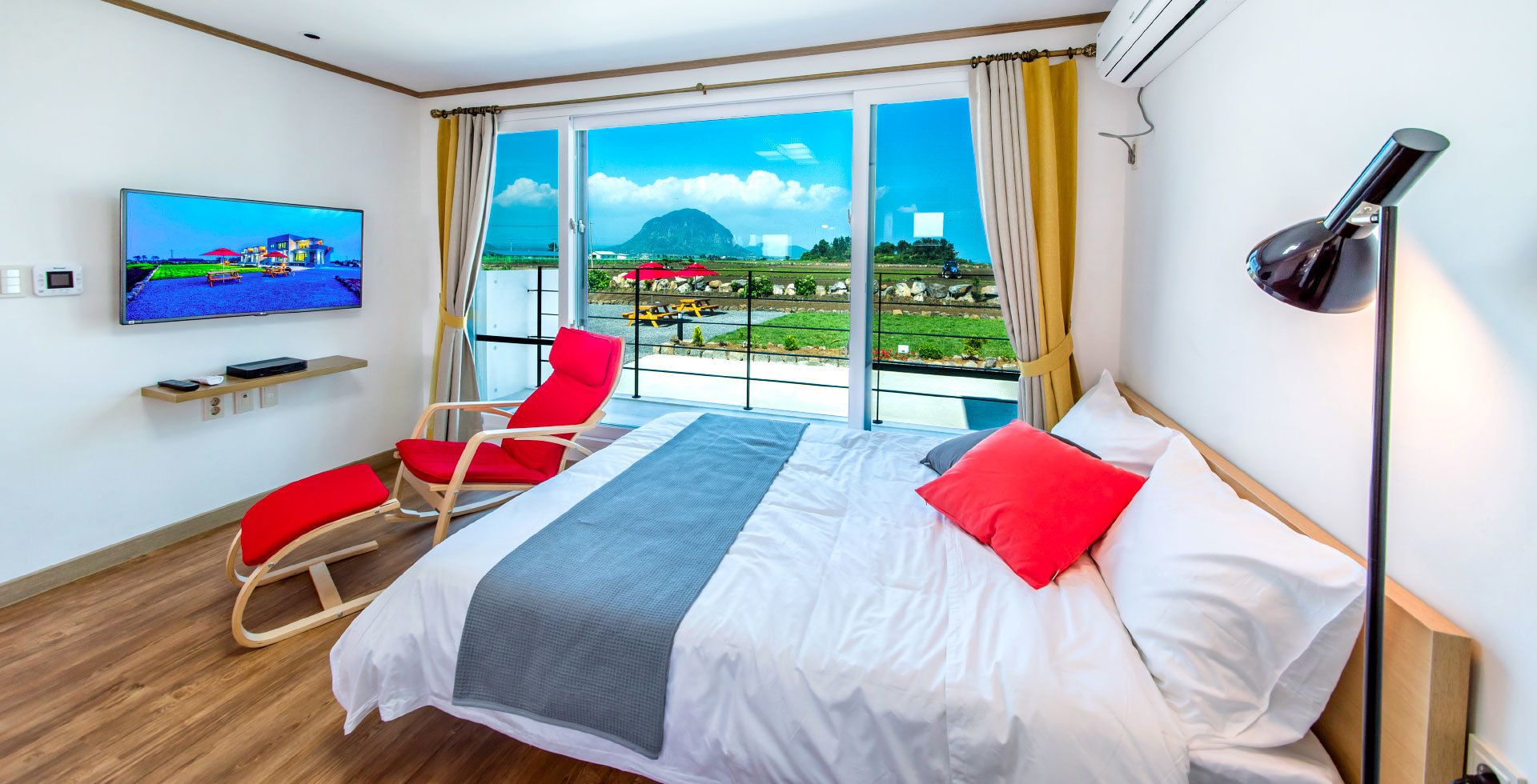 Une chambre d’hôtel avec un grand lit, deux fauteuils rouges, un téléviseur fixé au mur et une vue sur un paysage comprenant des montagnes et une étendue d’eau.