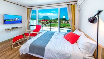 Habitación de hotel con una cama grande, dos sillones rojos, un televisor en la pared y vistas a un paisaje con montañas y una masa de agua.