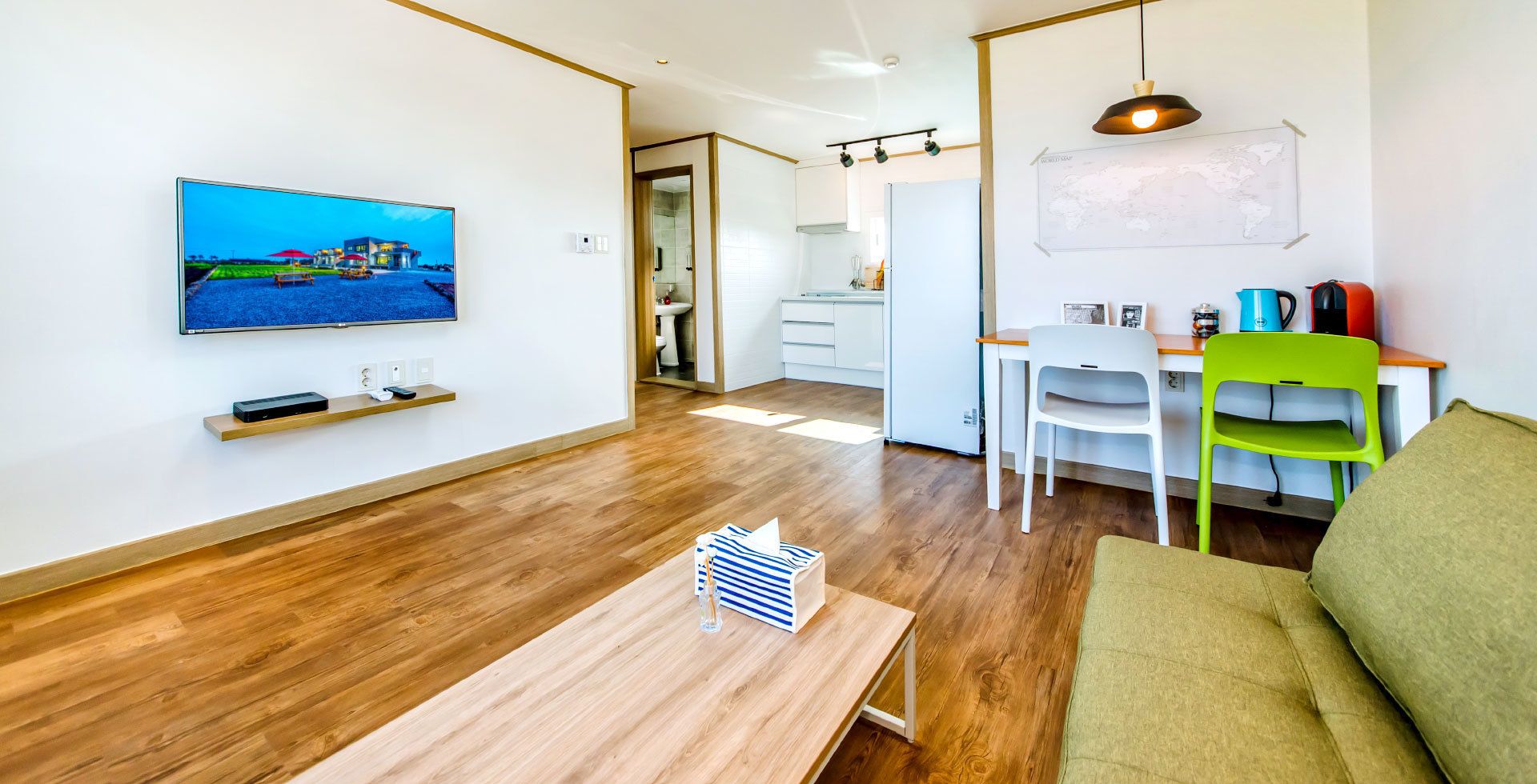 Un salón moderno con suelo de madera, un televisor de pantalla plana montado en la pared, una zona de comedor con una mesa y sillas blancas, y un sofá verde.