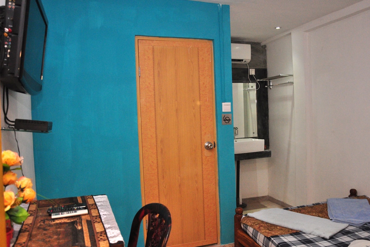 Una habitación con puerta de madera, una cama con colcha a cuadros, una mesita con un teclado y un espejo sobre la cama.