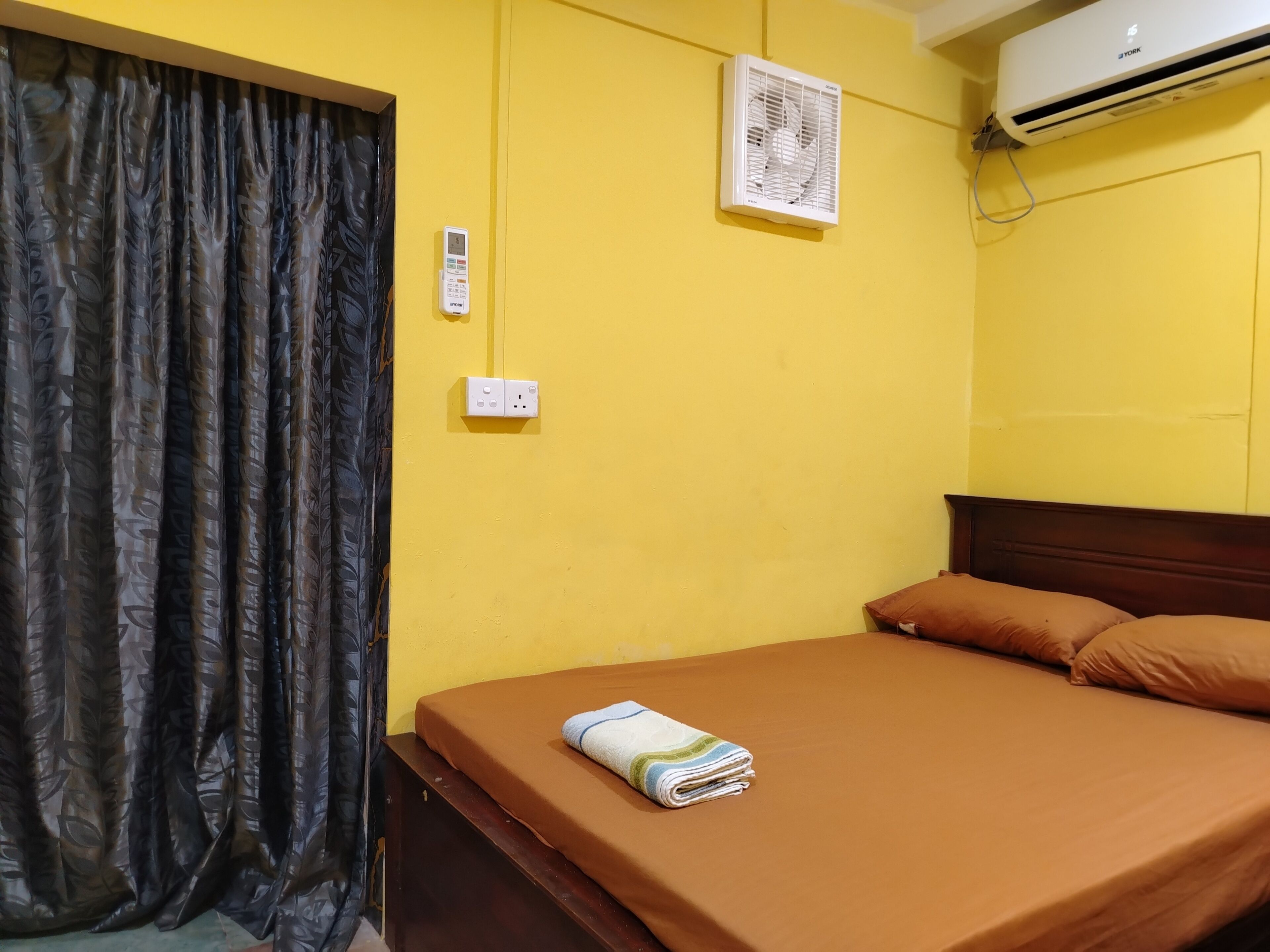 A room with a bed, a curtain, an air conditioner, and a wall socket.