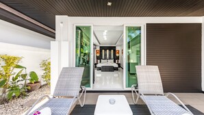 Un espacio lounge moderno al aire libre con dos sillones de mimbre, una pequeña mesa blanca y un ventanal de vidrio que da a un dormitorio con cama, mesitas de noche y un espejo.