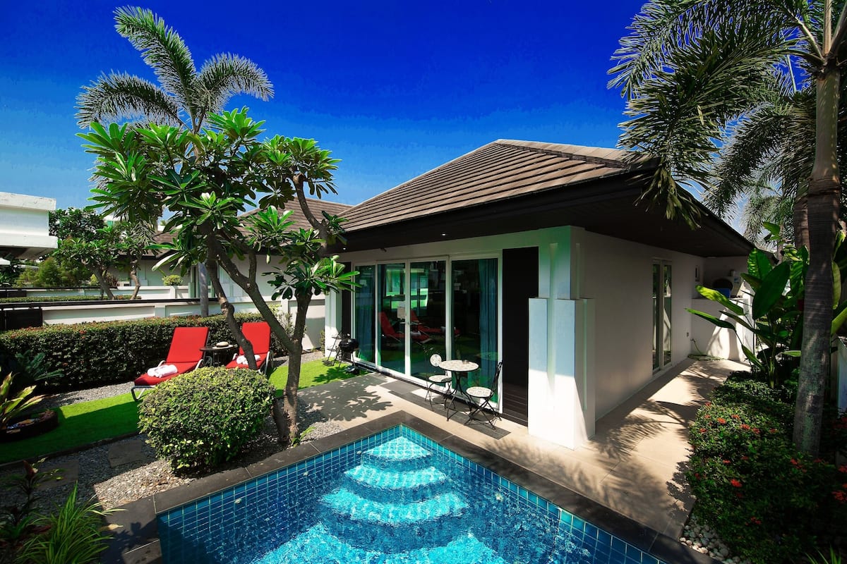 Ein modernes Haus mit Swimmingpool, roten Liegestühlen und tropischen Pflanzen.
