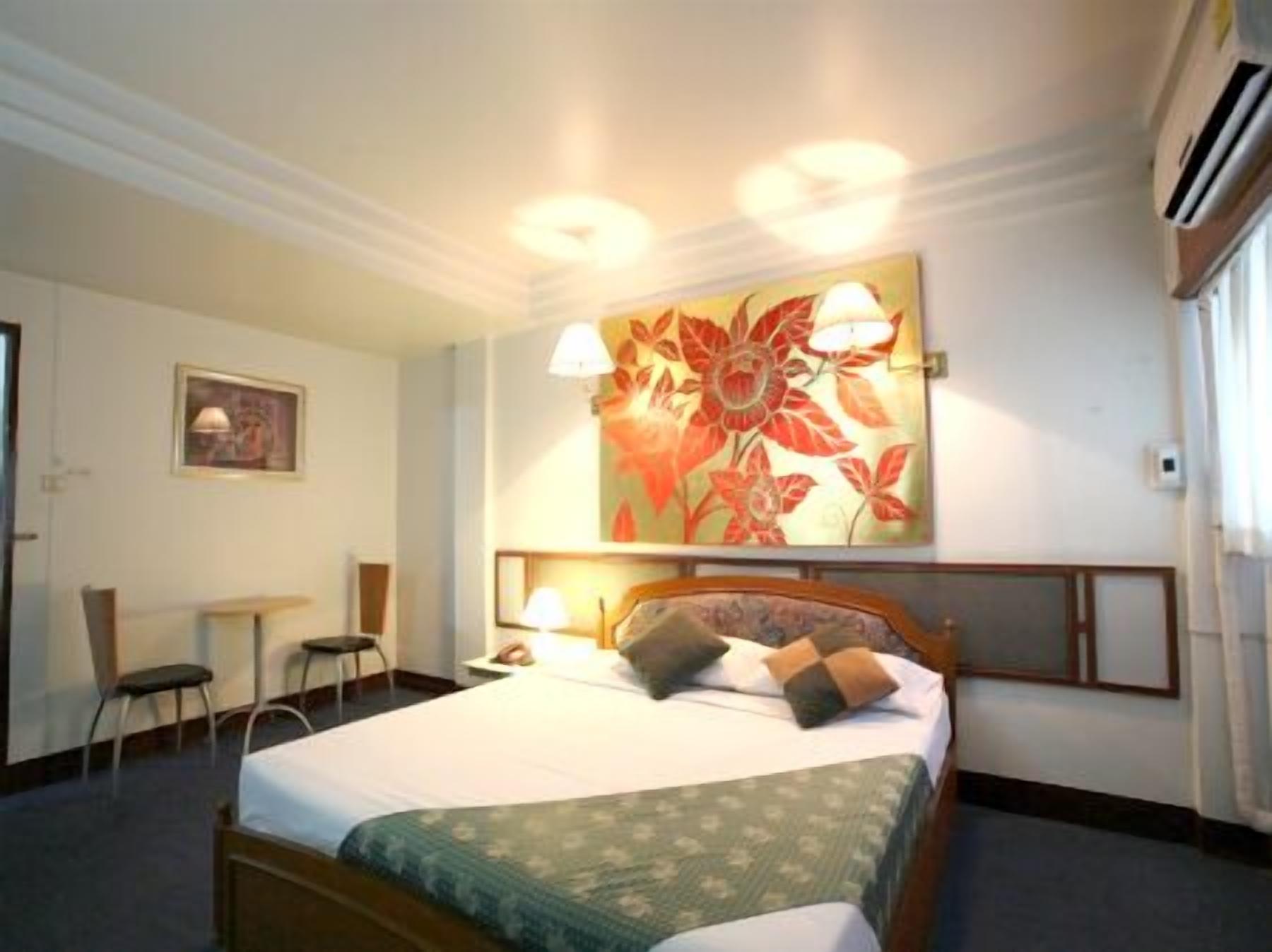 A hotel room with a bed, two bedside lamps, a floral artwork on the wall, a small table with a chair, and an air conditioning unit.