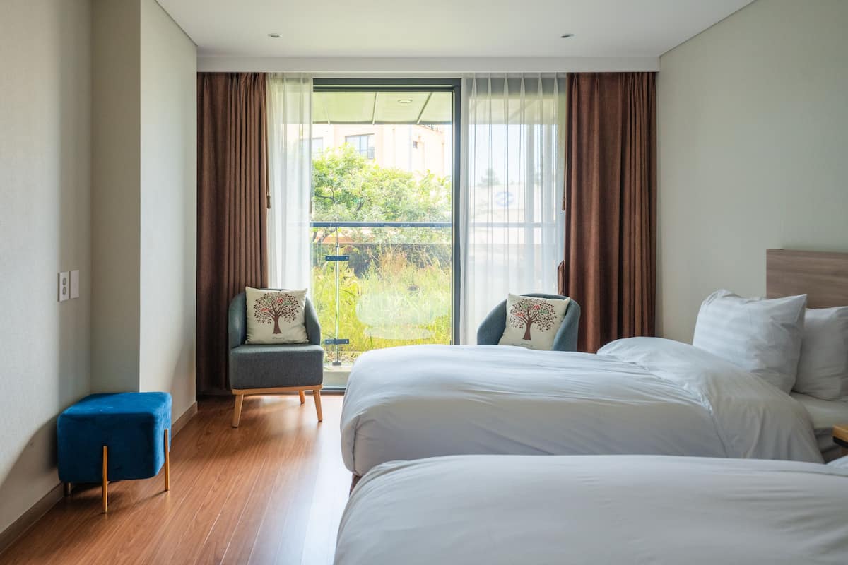 Hotellihuone, jossa on kaksi sänkyä, pieni sininen jalkarahi, kaksi nojatuolia ja parveke, jossa on verhot.