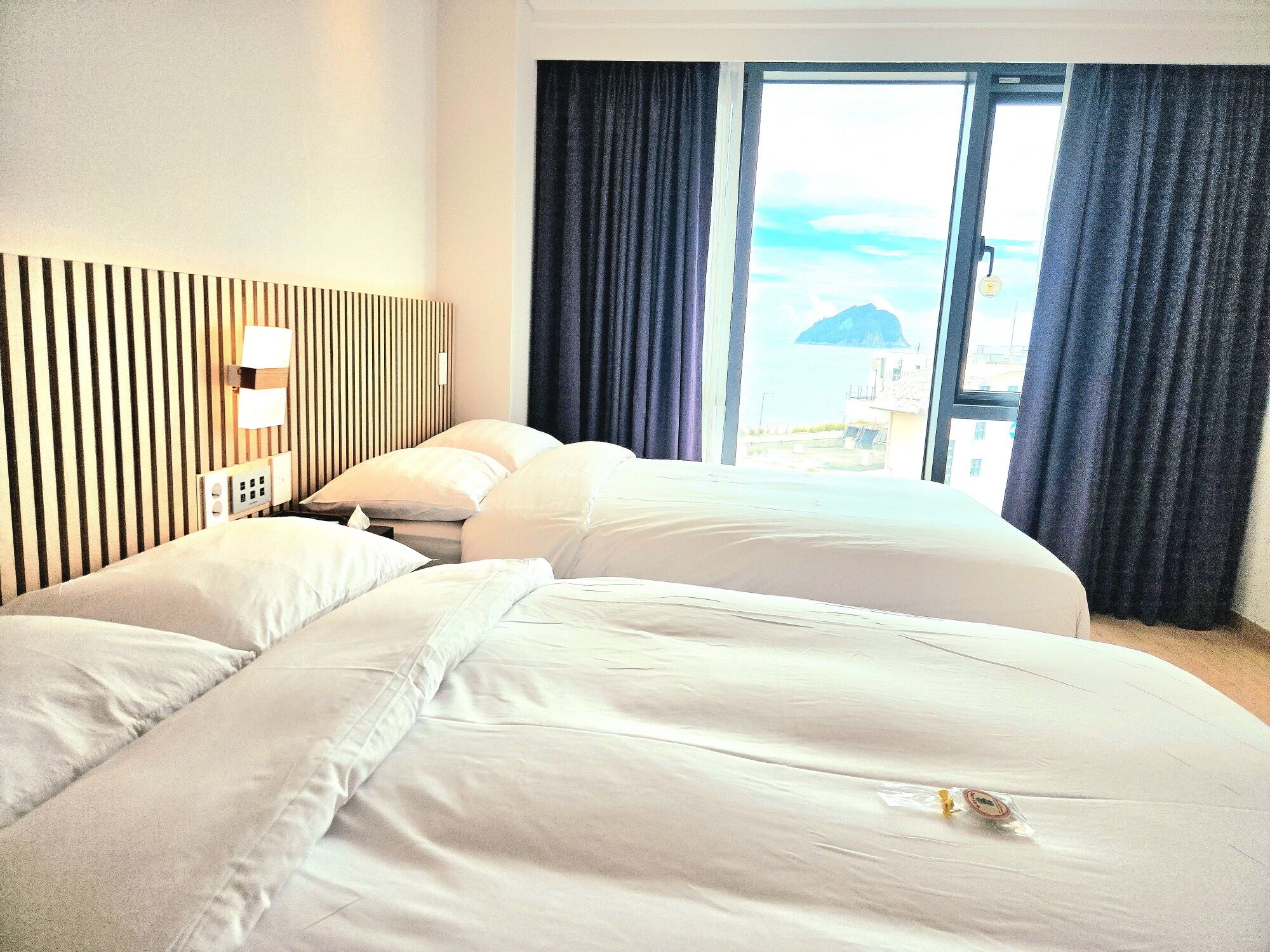 Habitación de hotel con dos camas, pared a rayas, ventana con cortinas y vistas a edificios.