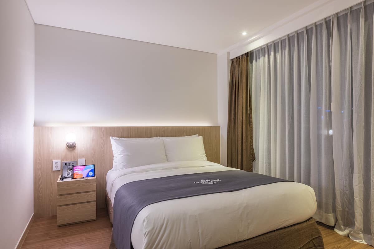 Moderni hotellihuone, jossa on siististi pedattu sänky, yöpöytä, jossa on kannettava tietokone, ja näkymä kaupunkiin ohuen verhon läpi.