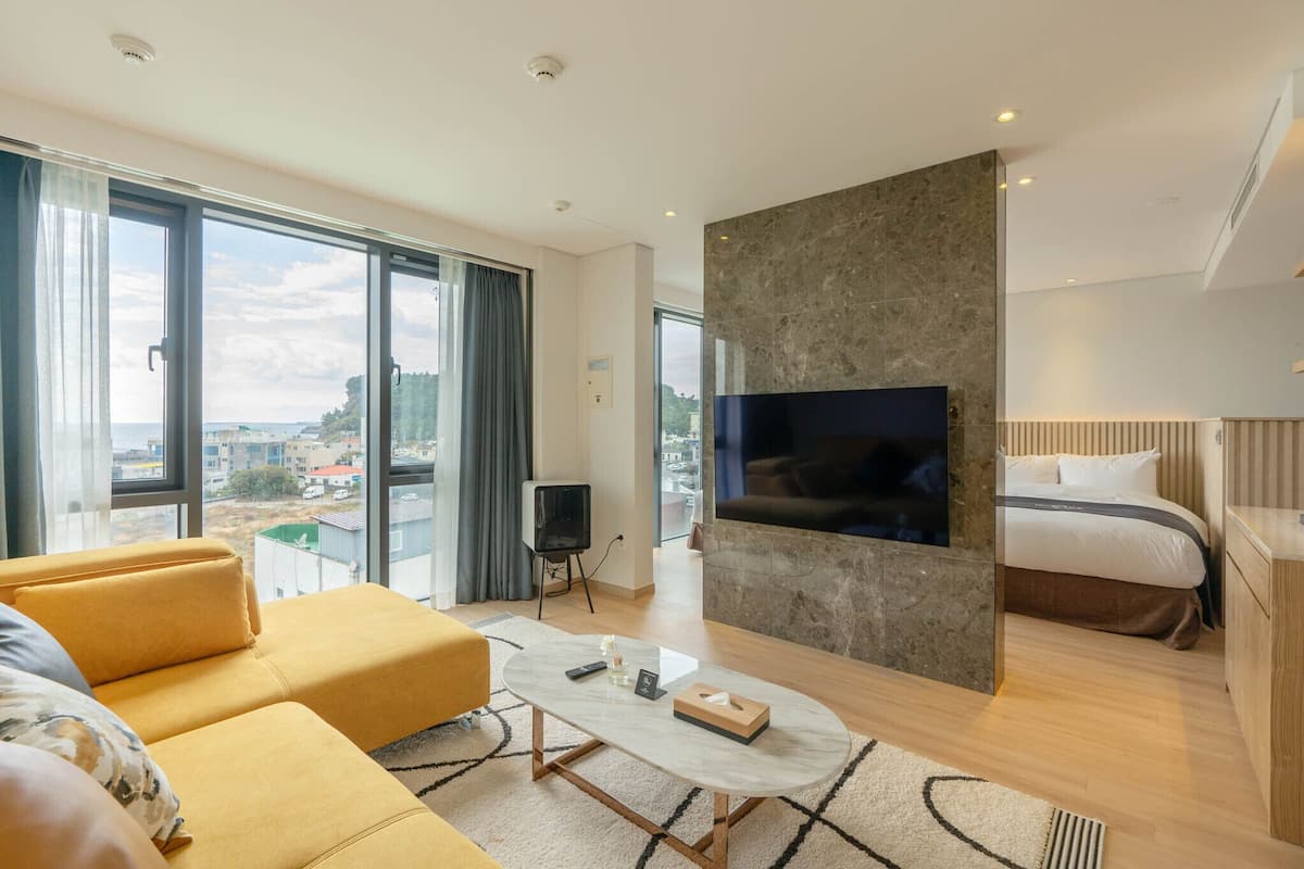Moderni hotellihuone, jossa on suuri sänky, sohva, kiviseinälle asennettu televisio ja suuret ikkunat, joista avautuu näkymä kaupunkiin.