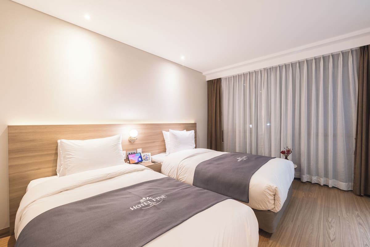 Hotellihuone, jossa on kaksi sänkyä, puinen pääty, yöpöytä lampun kanssa ja näkymä ulos verhojen läpi.