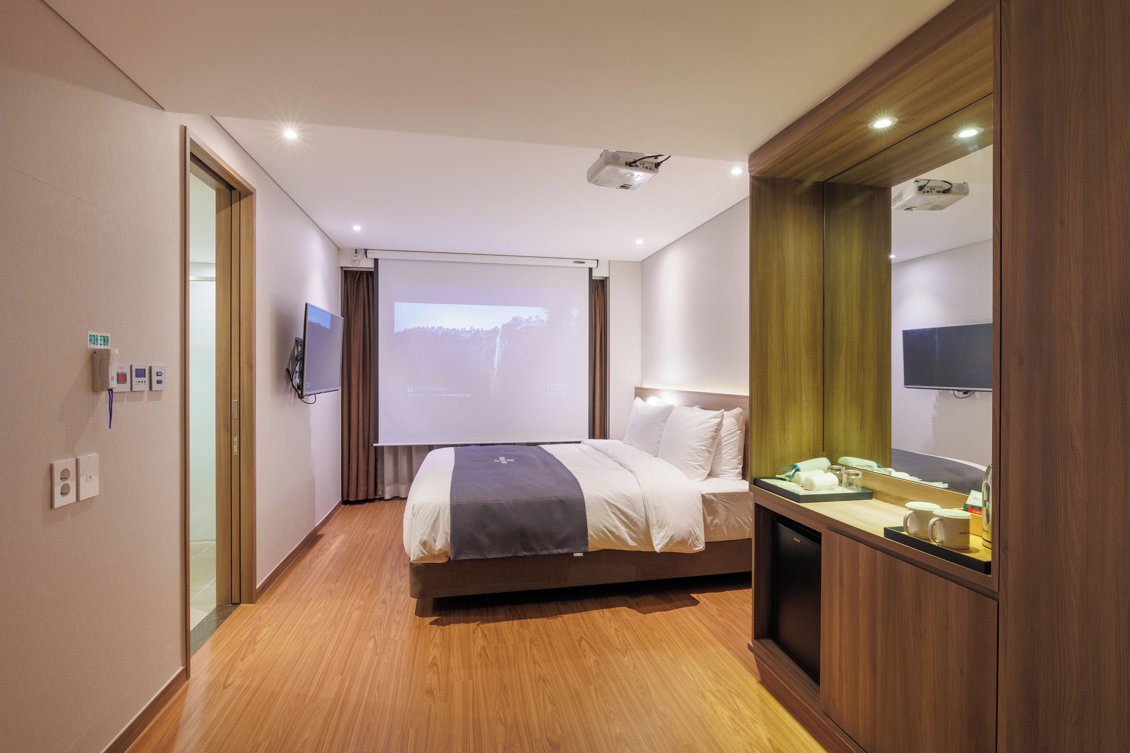 A hotel room with a bed, a desk with a telephone and a cup, a projector screen, and a television.