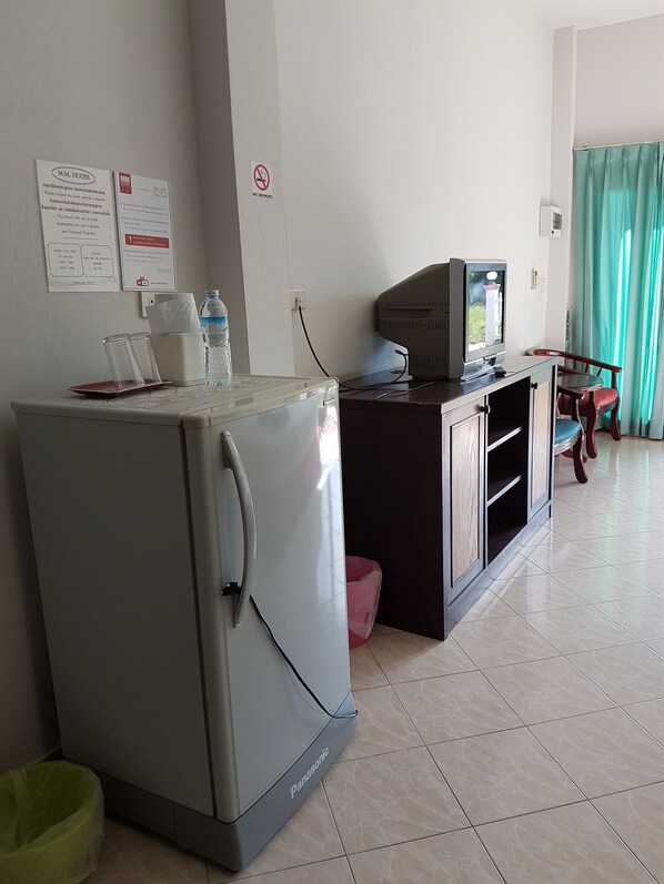 A compact kitchenette with a refrigerator, a microwave, and a small TV. There are signs on the wall, a trash bin, and a chair near a window with curtains. - MM House Samui (Koh Samui)