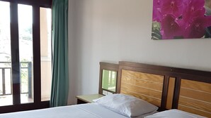 A neatly made bed with white linens and a folded towel, a wooden headboard, a nightstand with a lamp, and a painting of pink flowers on the wall. - MM House Samui (Koh Samui)