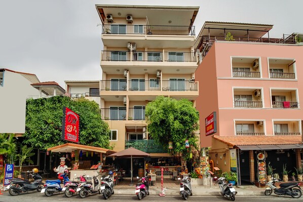 Un edifici de diversos pisos amb balcons, un restaurant amb terrassa i diverses motores aparcades.