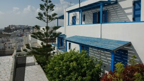 A white building with blue accents and a sloped roof, surrounded by greenery and other buildings in the background. - Ocean View (Naxos)
