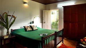 A bedroom with a green bedspread, wooden headboard, a nightstand with a vase and books, a wall-mounted light, and a door leading to a balcony with plants. - Harvest Fresh Farms (Uthamapalayam)