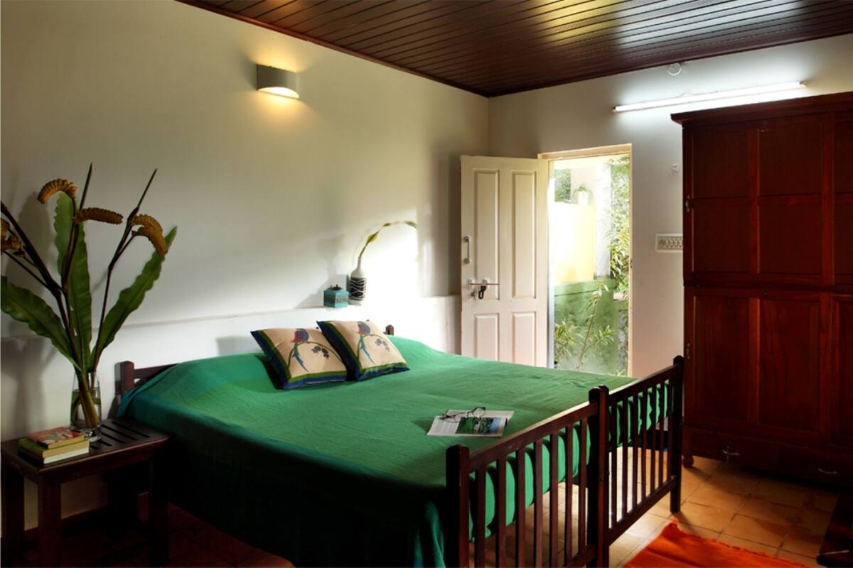 A bedroom with a green bedspread, wooden headboard, a nightstand with a vase and books, a wall-mounted light, and a door leading to a balcony with plants.