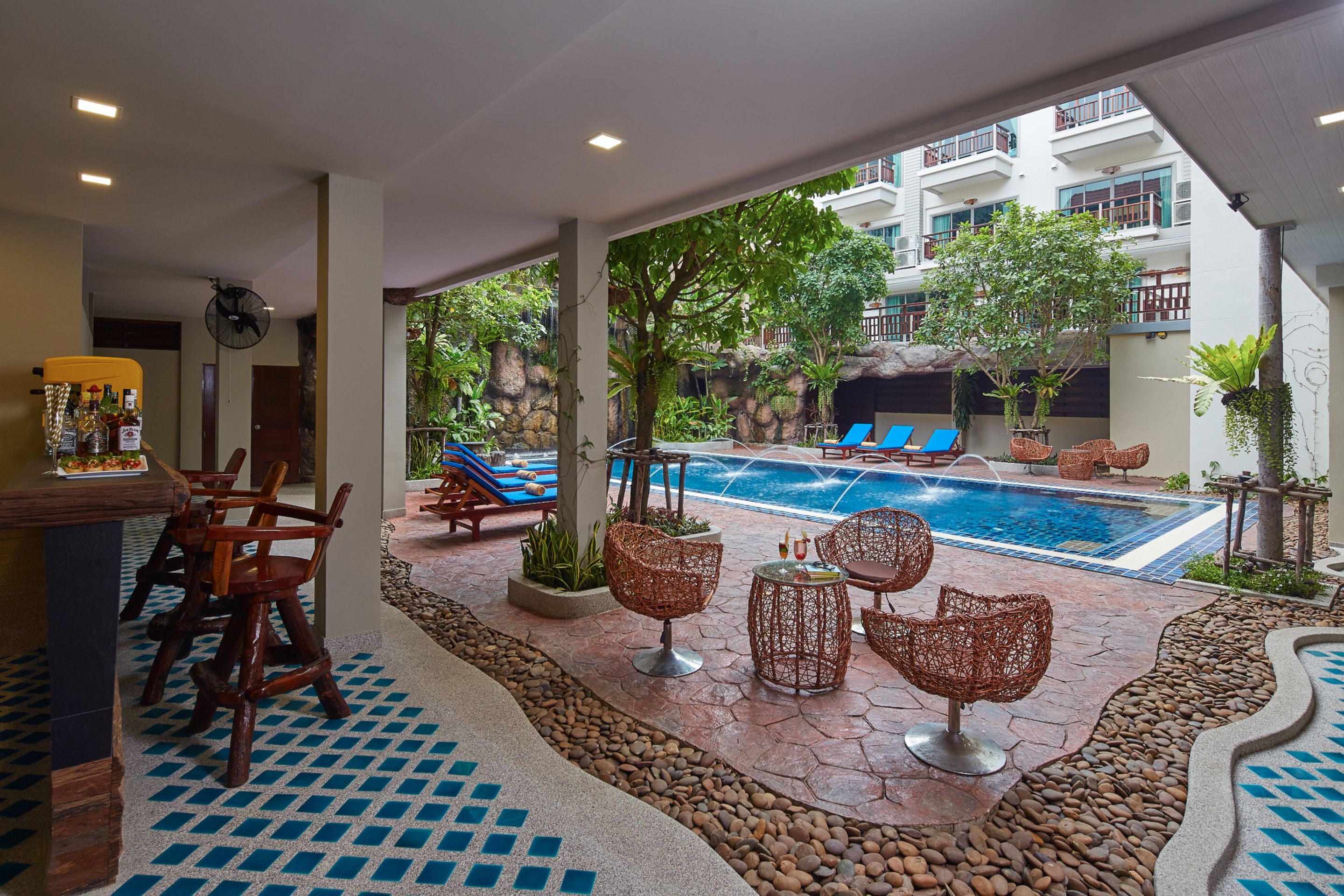 Zona de piscina con sillones reclinables, un bar y vistas a un edificio con balcones.