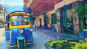 Un tuk-tuk est garé devant un immeuble dont la façade est en bois, et un grand bac à fleurs, rempli de plantes, se trouve à proximité.