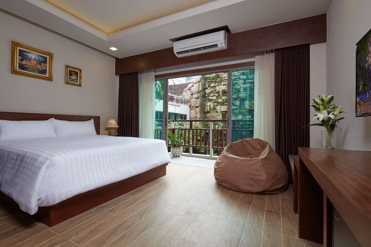 A hotel room with a large bed, a desk, a bean bag chair, and a balcony with a view of greenery.