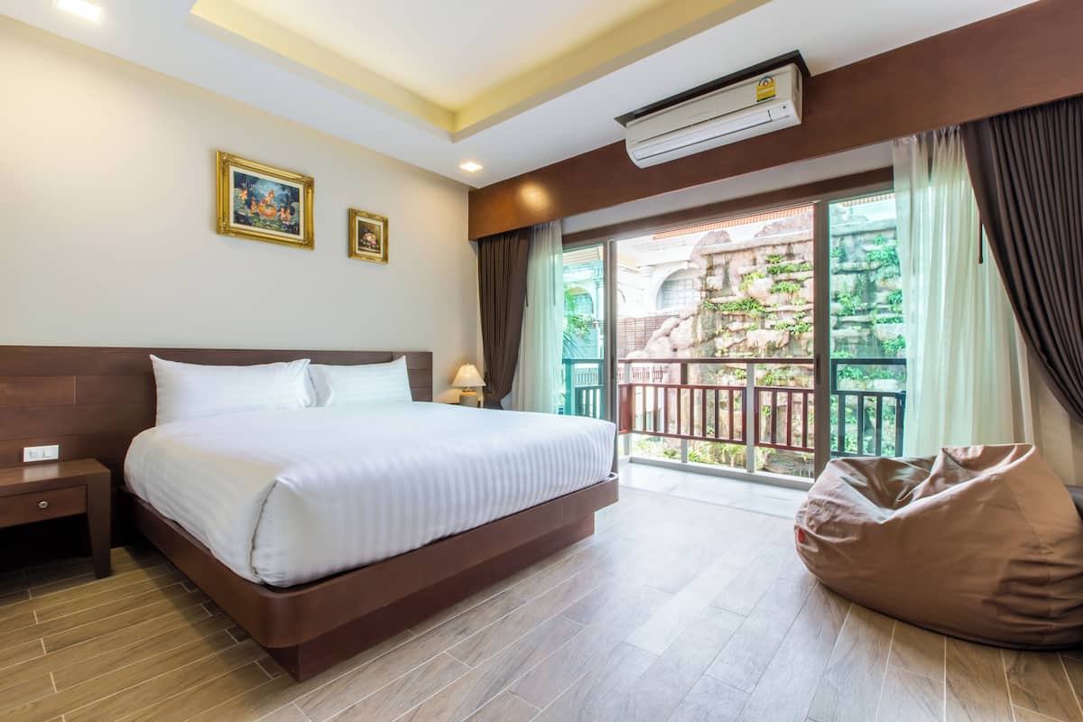 A hotel room with a large bed, a bedside table, a nightstand, a bean bag chair, and a balcony with a view of greenery.
