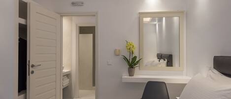 A hotel room with a bed, a desk, a chair, a mirror, and a potted plant.