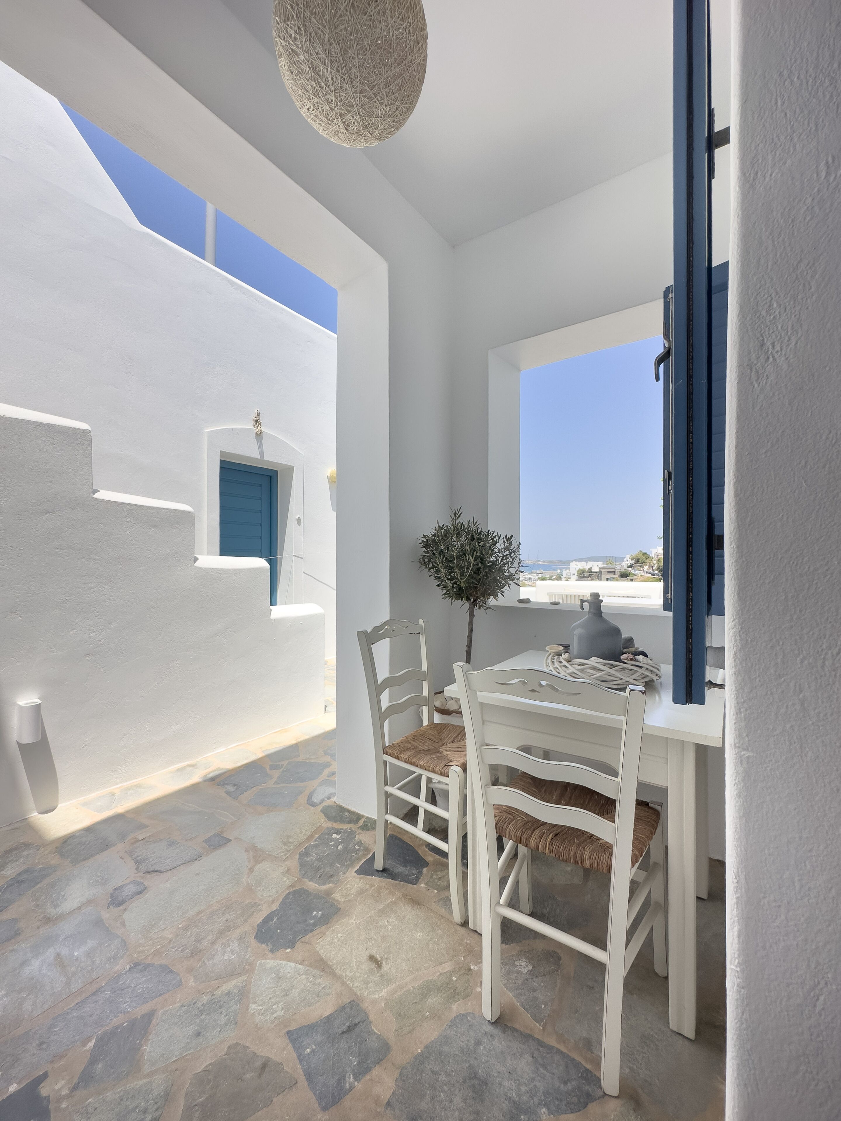 A white-walled room with a stone floor, a table with chairs, and a view of the sea.