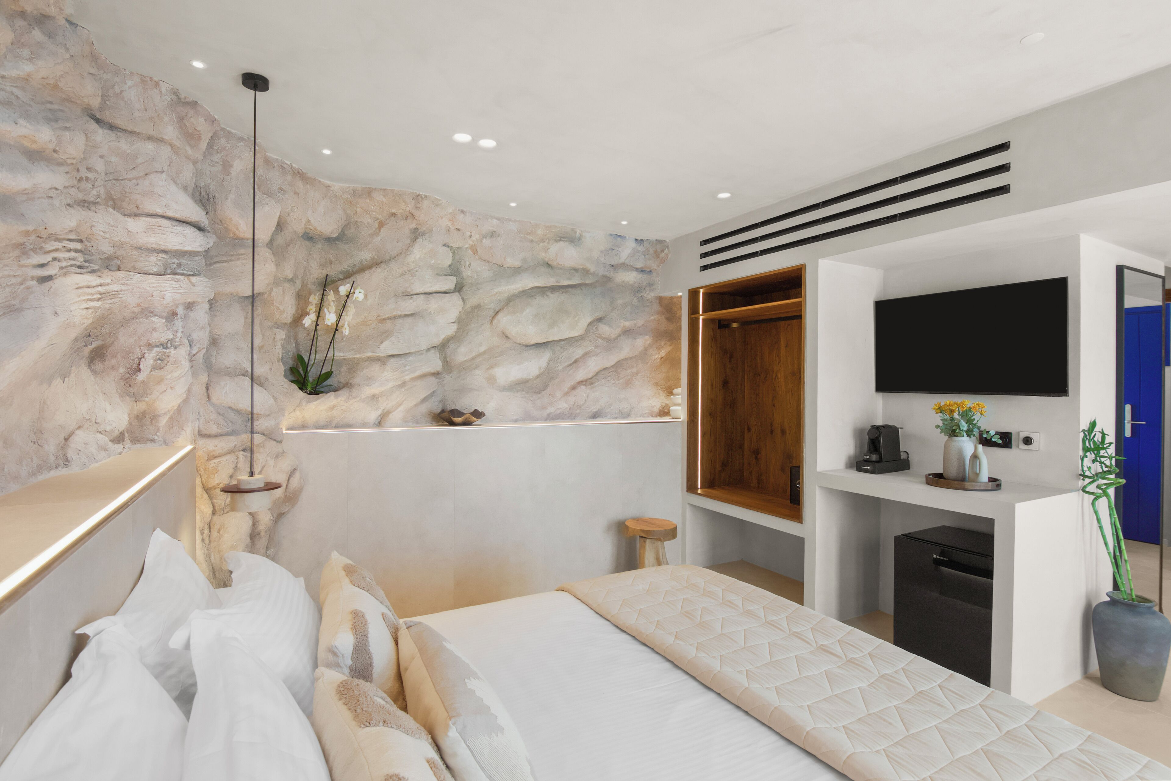 A modern bedroom with a stone wall, a bed with white bedding, a mounted TV, and a small desk area.