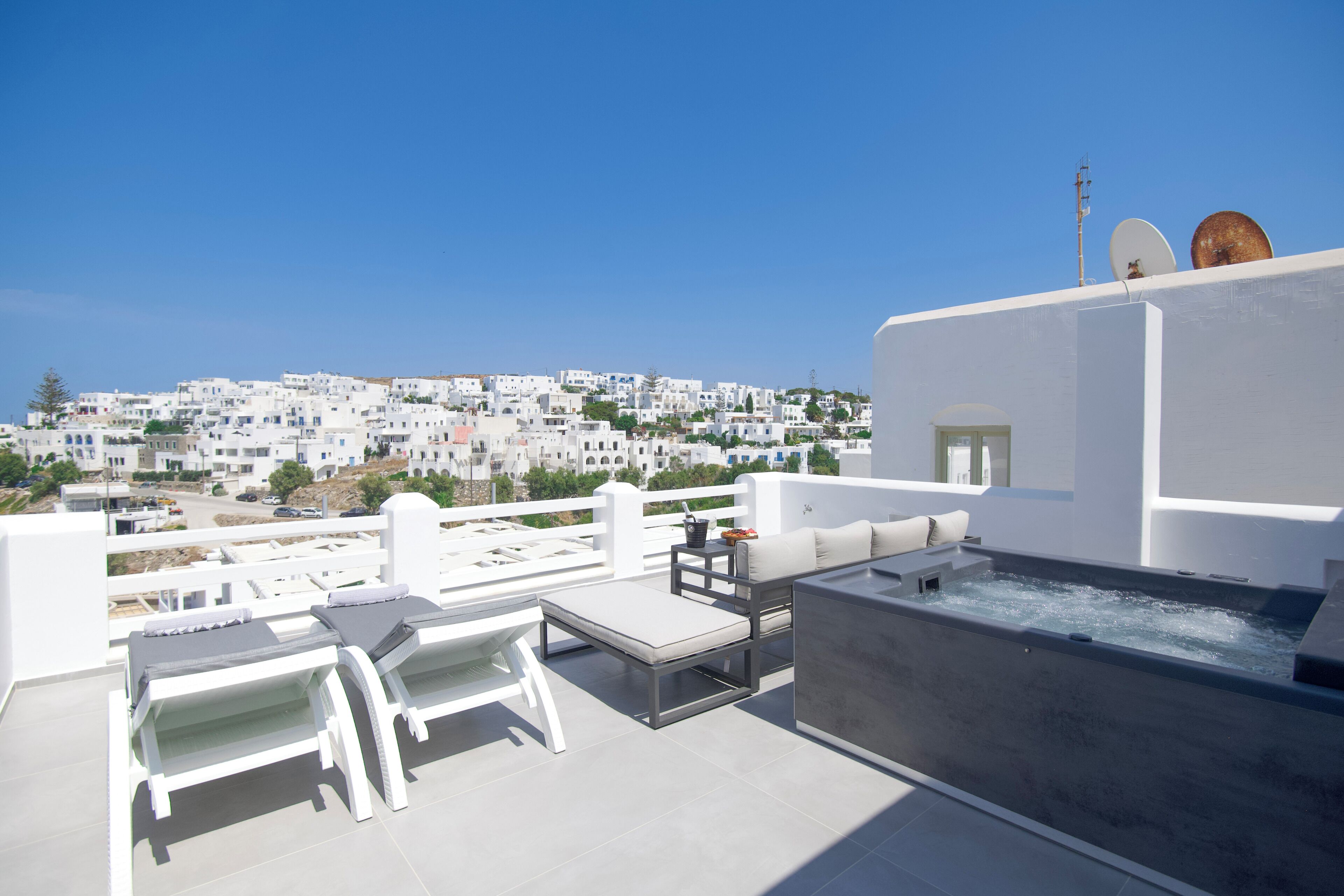 A rooftop terrace with white furniture, a hot tub, and a view of a white-washed town.