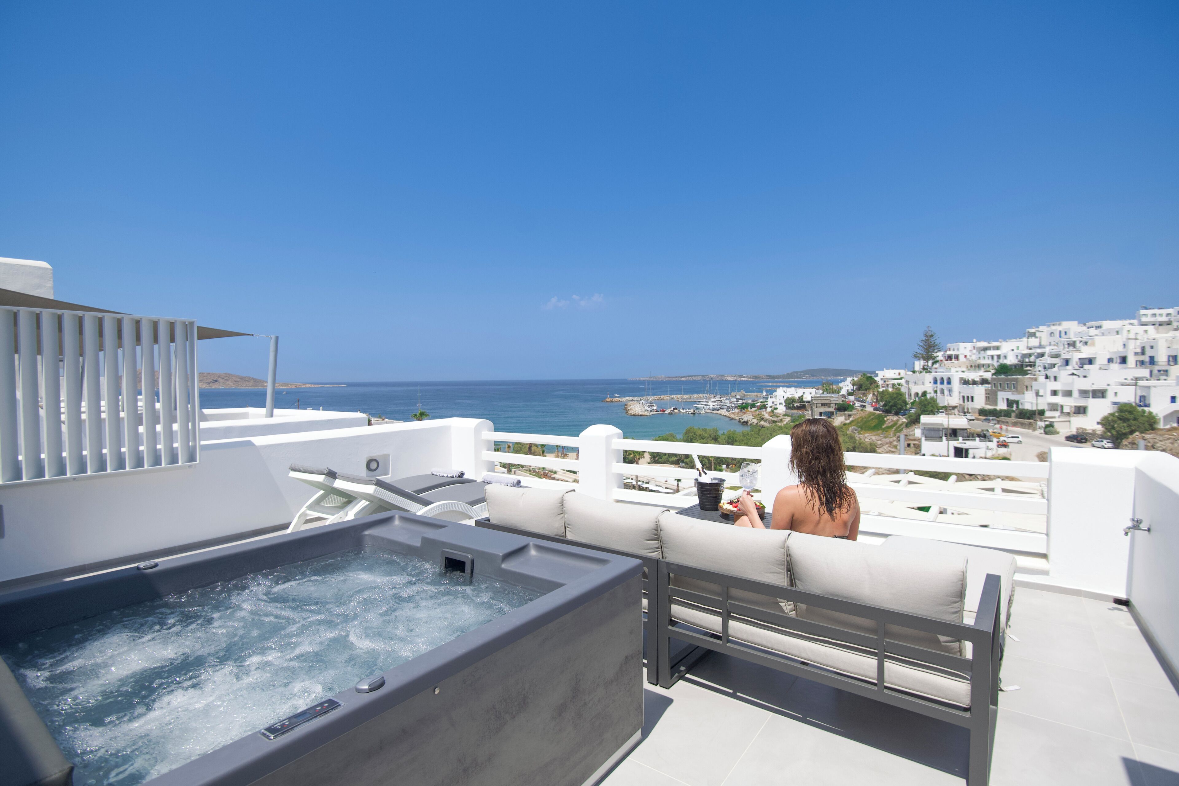 En balkon med jacuzzi, hvid gelænder og udsigt til havet og bygninger.