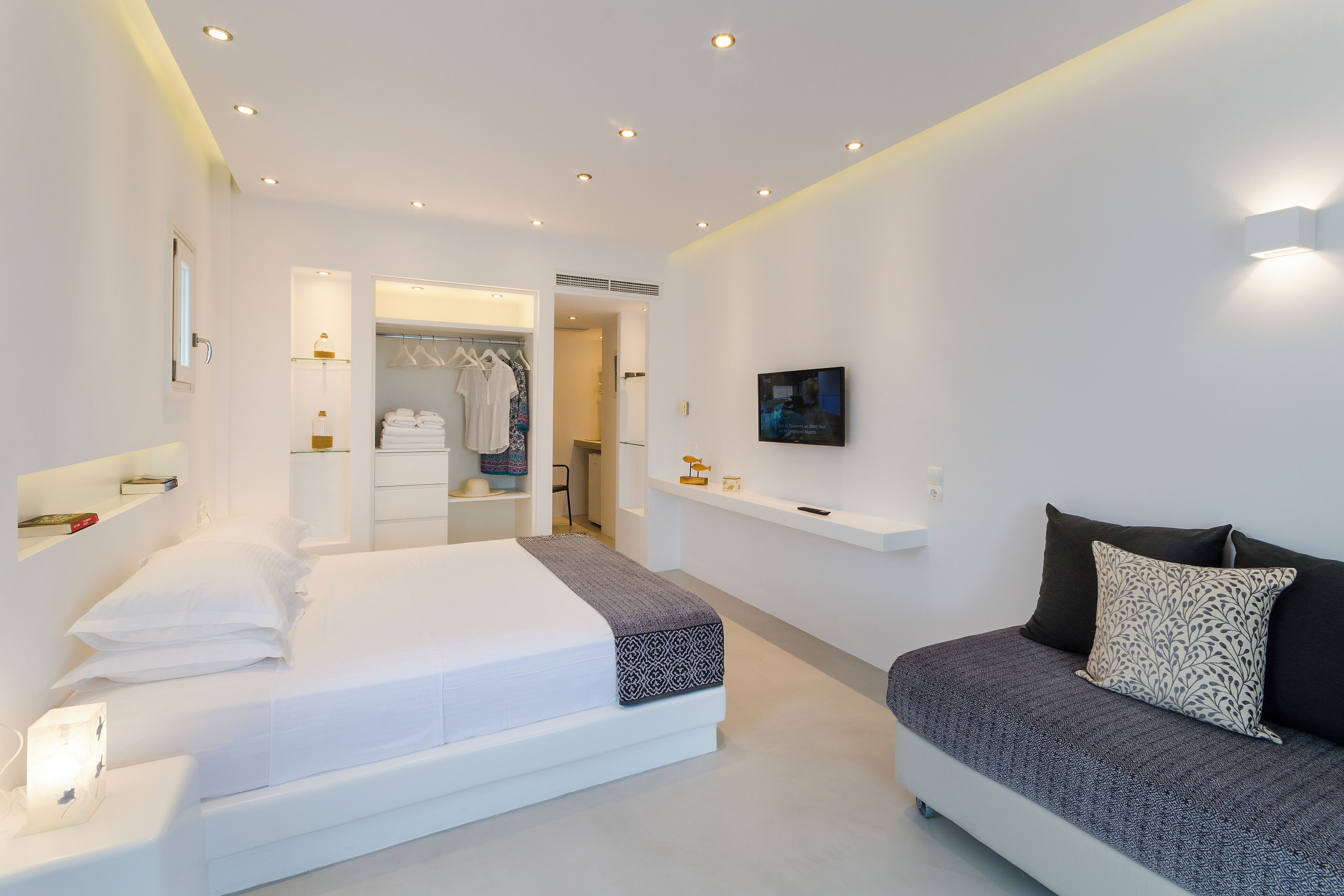 A modern bedroom with a large bed, a smaller sofa, a walk-in closet, and a TV on the wall.