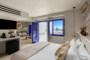 A modern bedroom with a large bed, a desk, and a view of the sea through a window.