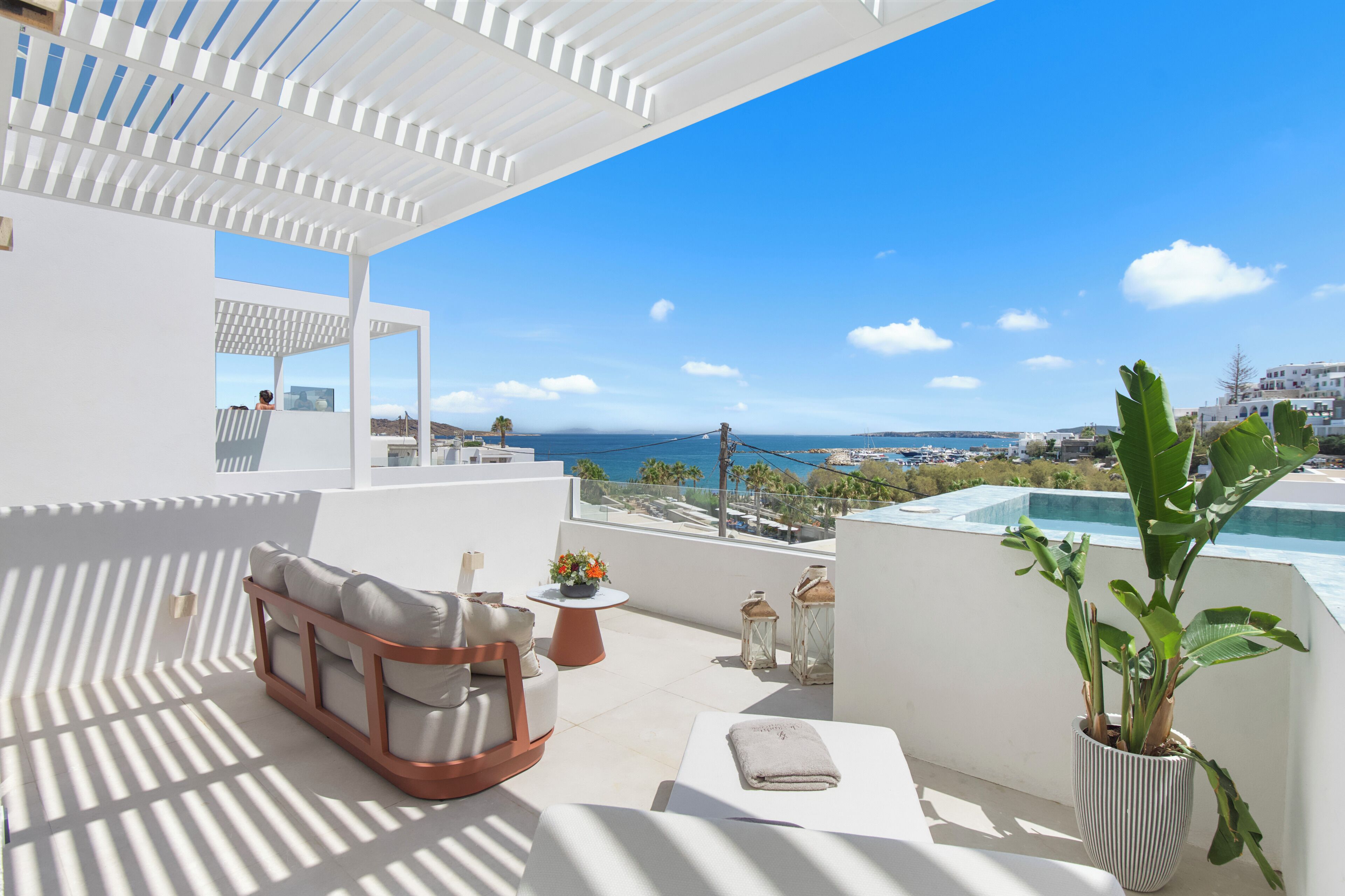 Une terrasse sur le toit, avec un espace extérieur aménagé de meubles blancs, une petite table avec un pot de fleurs, une plante en pot et une vue sur la mer et les bâtiments éloignés.