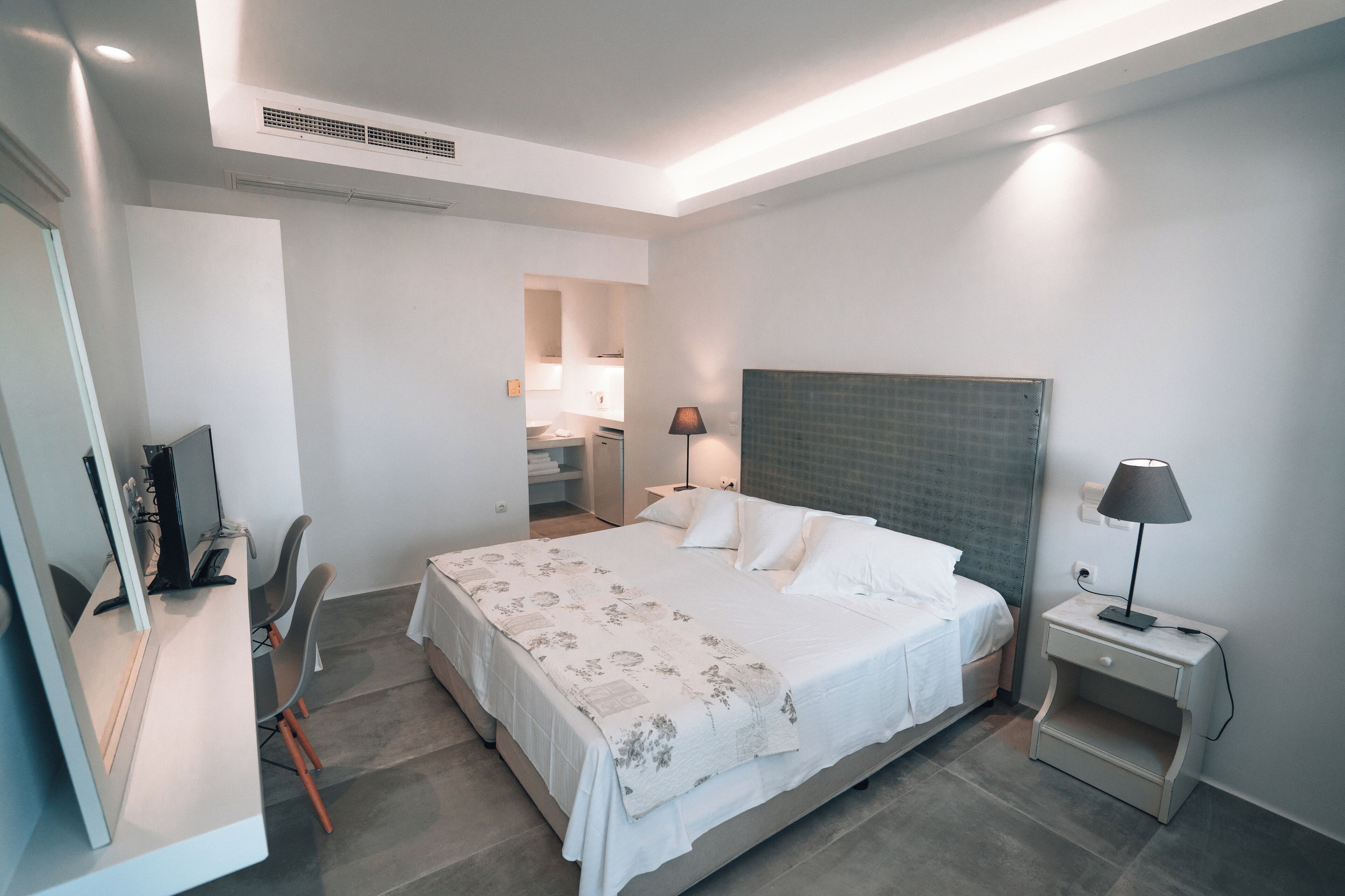 A modern hotel room with a large bed, bedside tables, a desk with a chair, and a bathroom visible in the background.