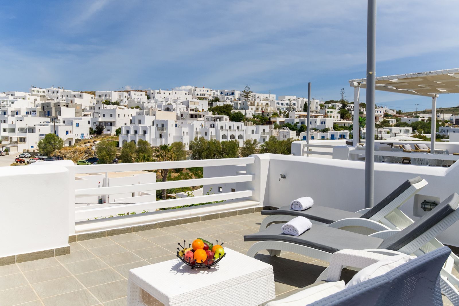 A rooftop terrace with white furniture, a fruit bowl, and a view of white buildings in the background.