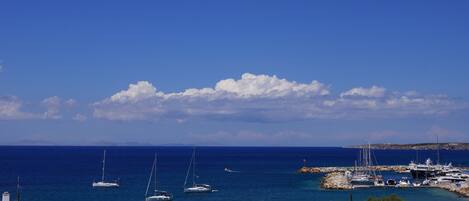 A coastal marina with several sailboats docked, a rocky breakwater, and a clear blue sky.