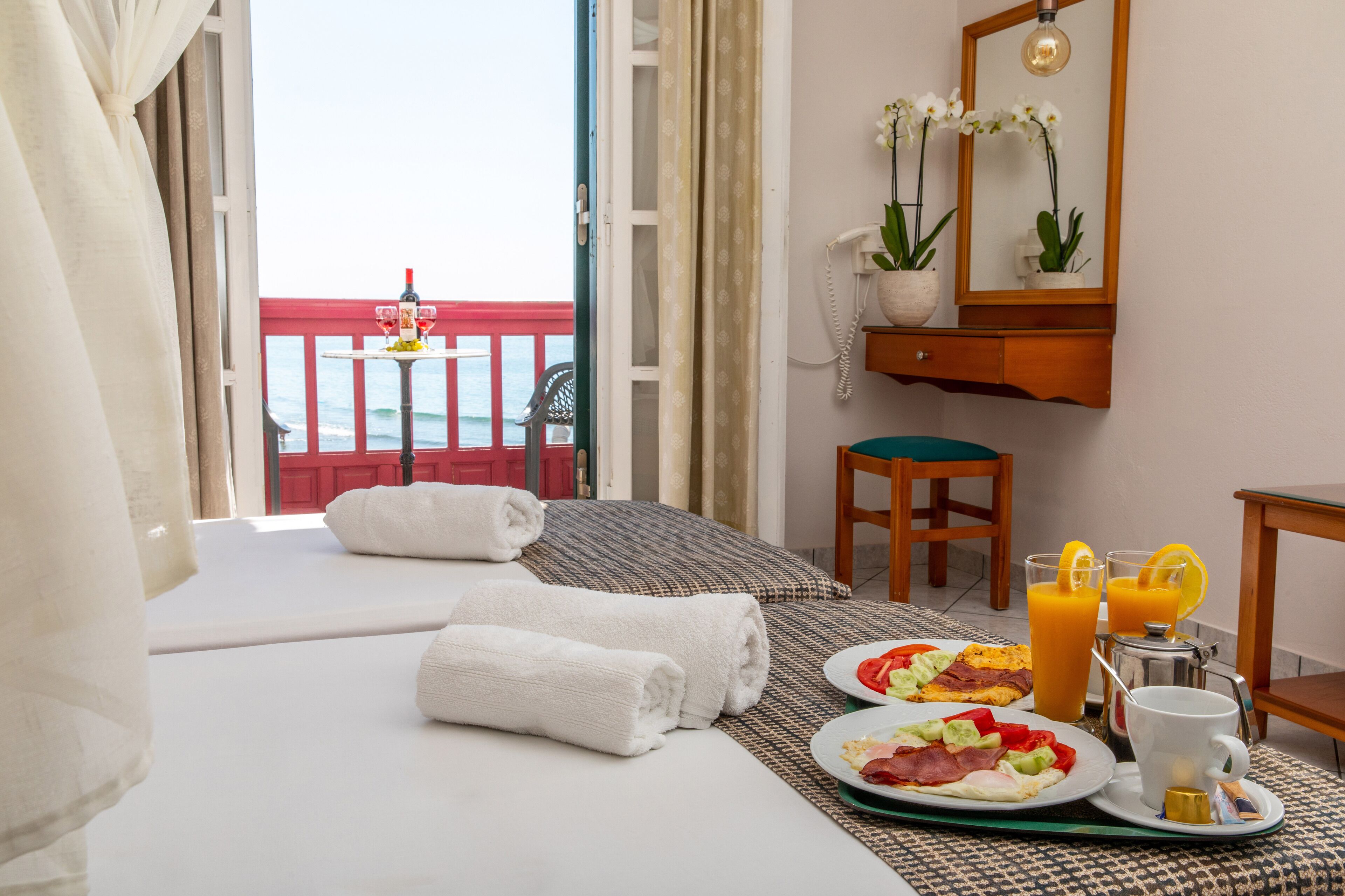 A hotel room with a bed, a breakfast tray with eggs, bacon, and orange juice, a coffee maker, and a view of the sea.