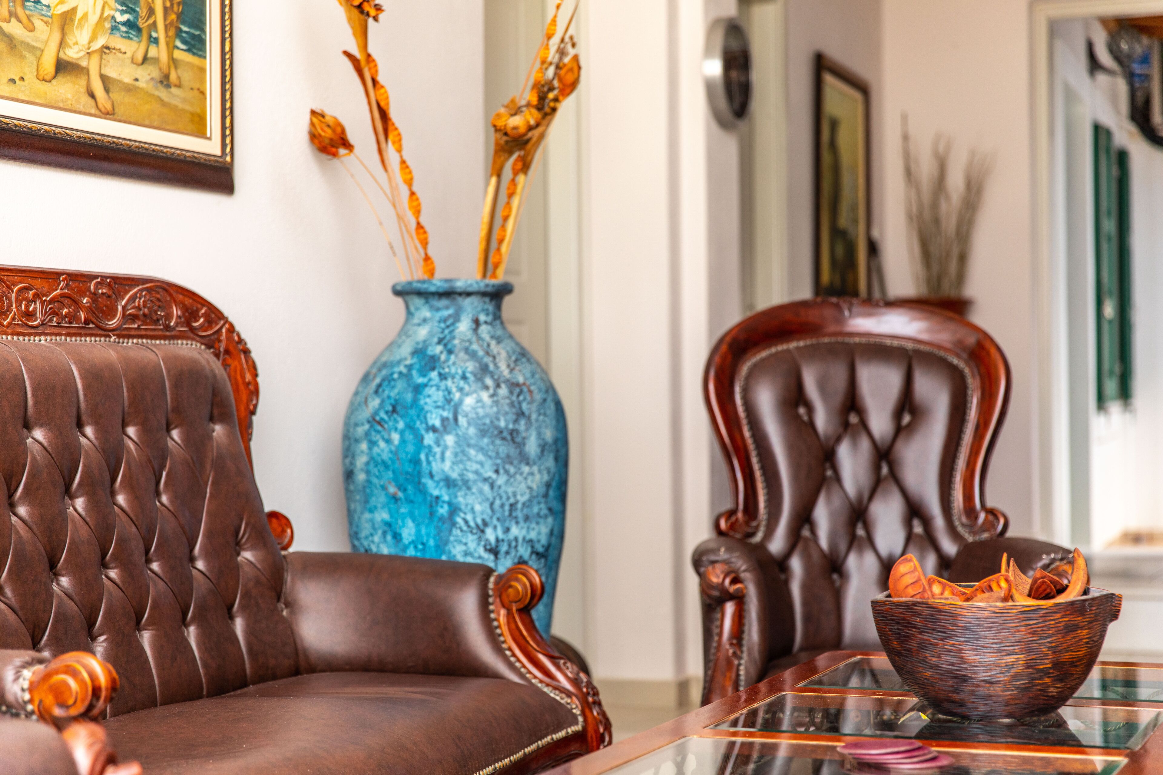 Una sala de estar con un sofá de cuero marrón, un jarrón azul con plantas secas, una silla de madera y una mesa de vidrio con una cesta tejida.