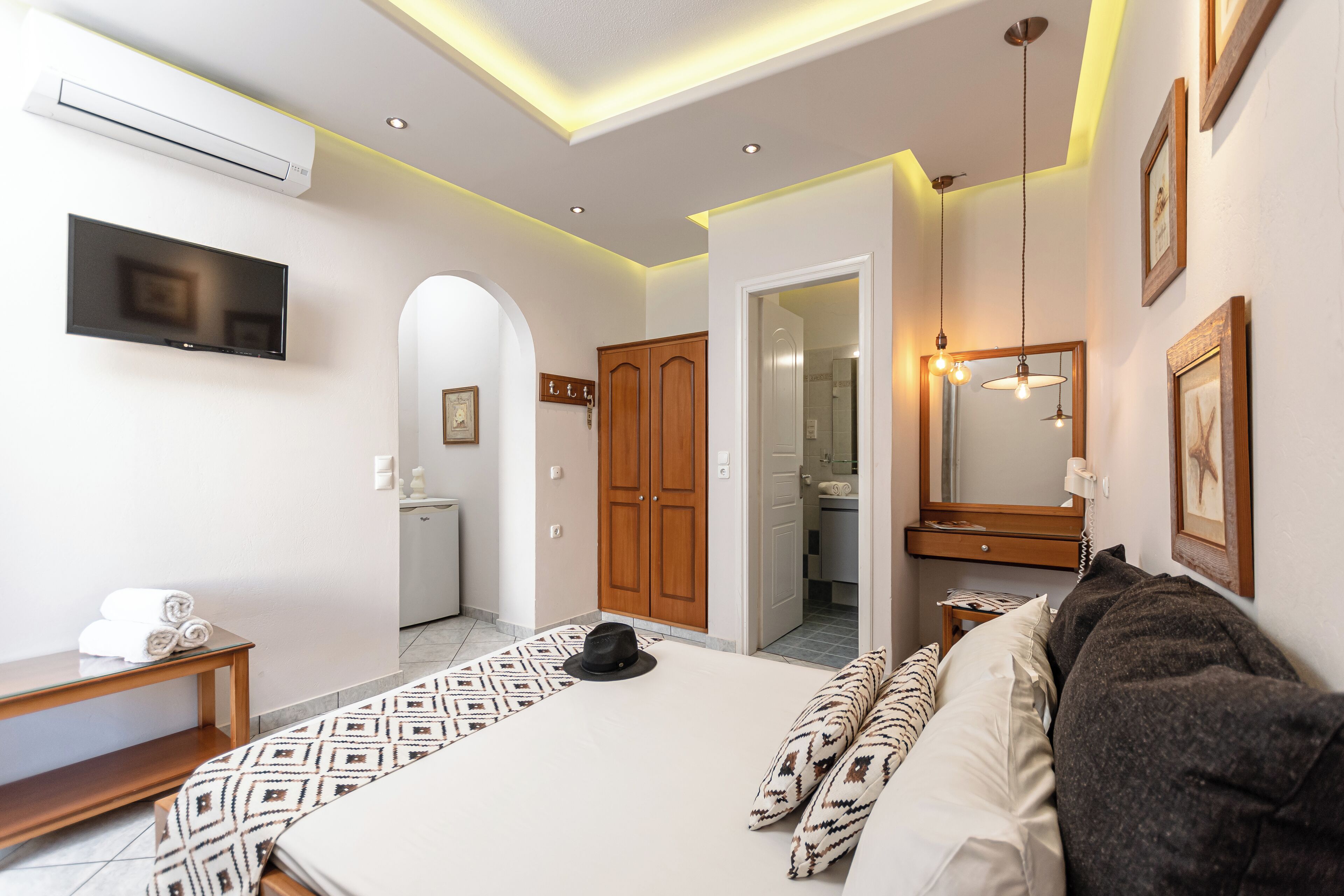 A modern hotel room with a large bed, a TV, a wardrobe, and a bathroom visible through an archway.
